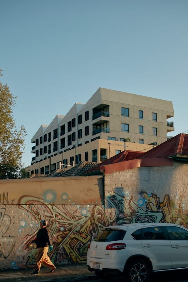 Exterior architectural photograph showing a contemporary multi-storey apartment building with a pale rendered or concrete facade, projecting balconies, dark-framed windows, and repeated angular roof forms. In the foreground are an older red-roofed structure, a graffiti-covered masonry wall, a pedestrian, and a parked car, creating an urban streetscape contrast.
