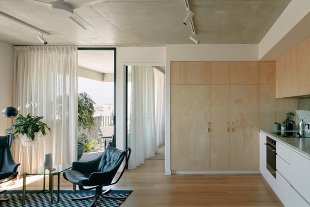 A bright open-plan interior combining a compact kitchen with a small living area. The room features floor-to-ceiling sheer curtains and glazing, a plywood-toned full-height built-in storage wall, white lower kitchen cabinetry, a dark stone-look benchtop, timber flooring, and a raw concrete ceiling. A black leather lounge chair, round side tables, indoor plants, and a patterned rug furnish the living zone. The overall aesthetic is restrained, contemporary, and warm with strong natural light.