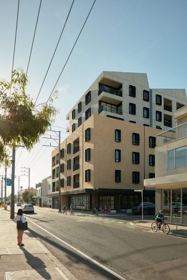 Urban streetscape showing a contemporary multi-storey apartment building with warm brick lower levels, pale rendered upper volumes, dark-framed windows, recessed balconies, and ground-floor commercial glazing. The building fronts a broad street with overhead utility wires, sidewalks, cyclists, pedestrians, and neighbouring low-rise development.