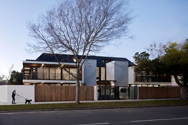 Street-facing exterior view of a contemporary multi-level residential building with a dark pitched roof, vertically ribbed light-metal facade panels, timber fencing, black metal gates and balcony railings. Large glazed openings and recessed warm lighting are visible, with a mature deciduous tree in front and a paved street in the foreground.