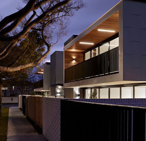 Twilight exterior view of a contemporary multi-storey residential building, with crisp white rendered volumes, dark metal balcony balustrades, expansive glazing, warm timber-lined soffits and integrated lighting. A brick boundary wall, timber fencing, paved walkway and mature deciduous trees frame the streetscape.