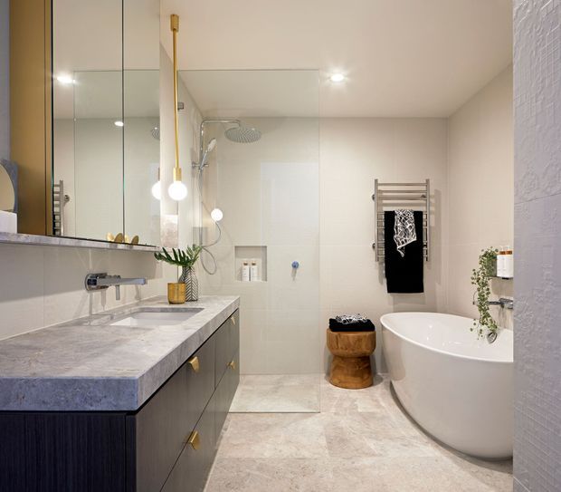 A bright, refined contemporary bathroom with a double vanity, large mirrored wall cabinet, walk-in glass shower, freestanding oval bathtub, heated towel rail and integrated storage. The room uses pale stone-look surfaces, dark timber-toned vanity cabinetry, brushed metal fixtures and warm brass accents.