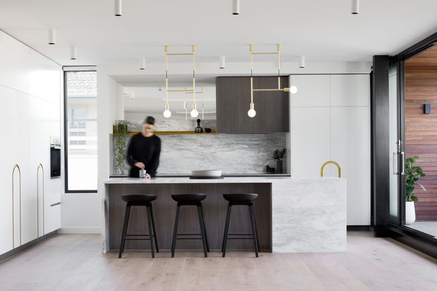 Bright contemporary kitchen with a large stone island, three dark timber stools, white integrated cabinetry, dark wood overhead joinery, and a marble-look splashback. Linear pendant lights and slim recessed ceiling lights create a clean, refined interior, with timber cladding visible through the full-height glazing at right.