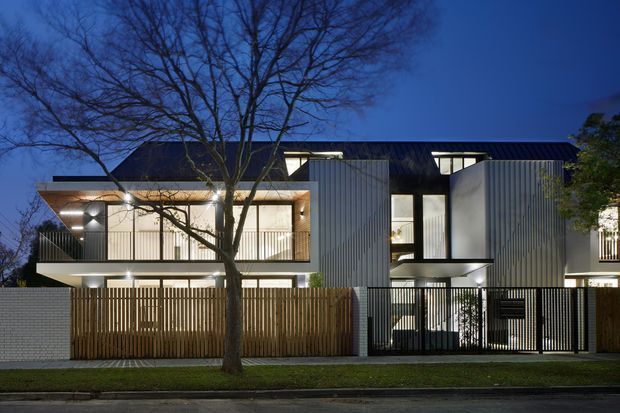 Twilight exterior view of a contemporary multi-storey residential building with a dark low-pitched roof, contrasting vertical corrugated metal cladding, pale masonry walls, warm timber-lined balconies and screens, and extensive illuminated glazing. A landscaped verge, mature street tree, timber privacy fencing, and metal gates occupy the foreground.