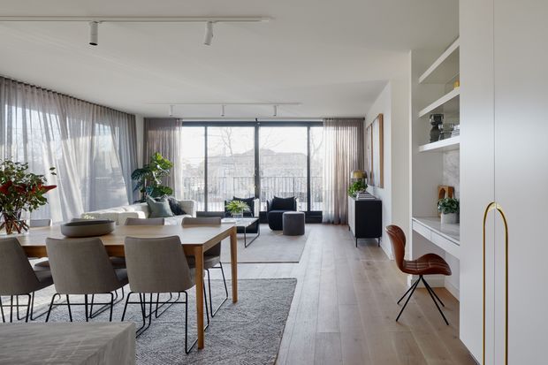 Bright open-plan living and dining room with floor-to-ceiling glazing, sheer curtains, pale timber flooring, a timber dining table with upholstered chairs, a compact dark sofa and contemporary built-in shelving. The space has a clean, restrained modern fit-out with track lighting and abundant natural light.