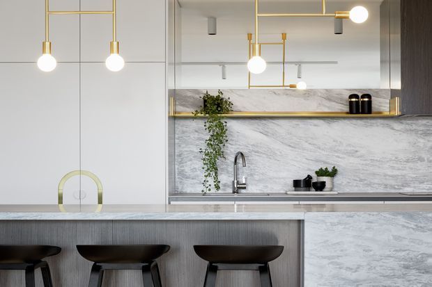 Contemporary kitchen interior featuring a large island with three dark sculptural bar stools, grey wood-grain cabinetry, a pale marble-look splashback and benchtop, and a reflective mirrored wall with slim brass shelving. Geometric brass pendant lights and restrained greenery add refined decorative detail to the light, polished space.