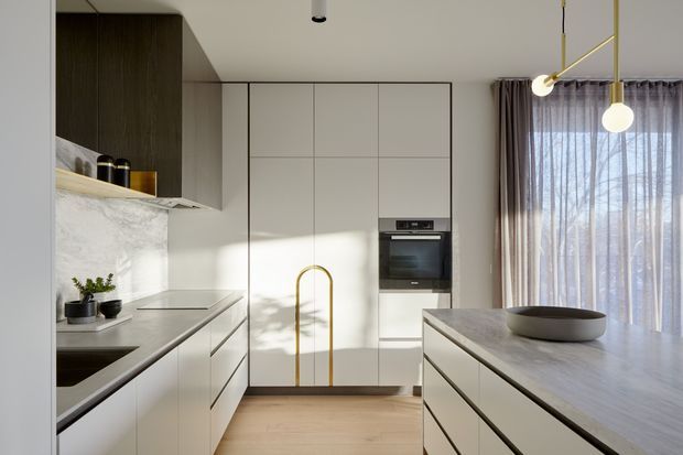 A refined contemporary kitchen viewed from the main work area, with full-height pale cabinetry, integrated oven, stone benchtops and splashback, a dark timber-toned overhead cabinet zone, and a large central island. A sculptural brass-toned tap, brass pendant lighting, sheer curtains, and light timber flooring add warmth. The kitchen has a clean, minimalist composition with abundant natural light.