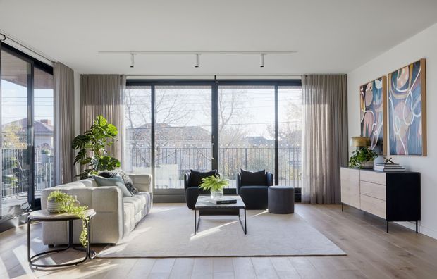 Bright contemporary living room with large floor-to-ceiling glazing, sheer curtains, a pale sectional sofa, dark lounge chairs, a low coffee table, and a streamlined sideboard beneath colourful abstract artwork. Natural light, indoor plants, and views of the surrounding neighbourhood create an airy, refined atmosphere.
