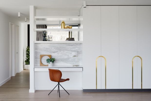 Bright contemporary kitchen and living entry area with full-height white cabinetry, distinctive brass loop handles, an open shelving niche, and a pale marble-look backsplash. A compact built-in counter with a sculptural brown swivel chair sits beside a hallway, with light timber flooring and minimal white surfaces throughout.