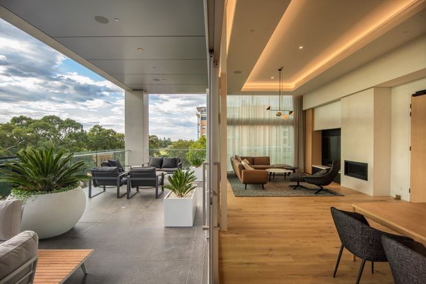 A contemporary open-plan living and dining area opening onto a covered balcony terrace through expansive glazing. The interior features warm timber flooring, a pale built-in fireplace wall, recessed cove lighting, sheer curtains, mid-century-inspired lounge chairs, and a dining setting. The balcony has grey tiled flooring, outdoor seating, and large white planters, with leafy suburban views beyond.