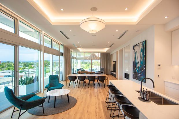 A bright, spacious open-plan kitchen, dining and living area with floor-to-ceiling glazing and expansive views. The room features light timber flooring, a large white island with seating, streamlined white cabinetry, a dining table with dark chairs, teal lounge chairs, layered recessed and cove lighting, and a contemporary circular pendant. A large abstract artwork and curtains add visual contrast.