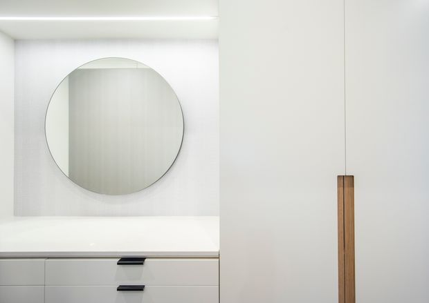 Minimalist close-up of a bright built-in vanity or storage joinery installation. White flat-panel cabinetry and drawers occupy the lower portion, with slim dark handles and a tall timber pull on the adjacent white cupboard. A large round mirror is mounted above the white benchtop, with concealed linear lighting creating an evenly illuminated, clean-lined composition.