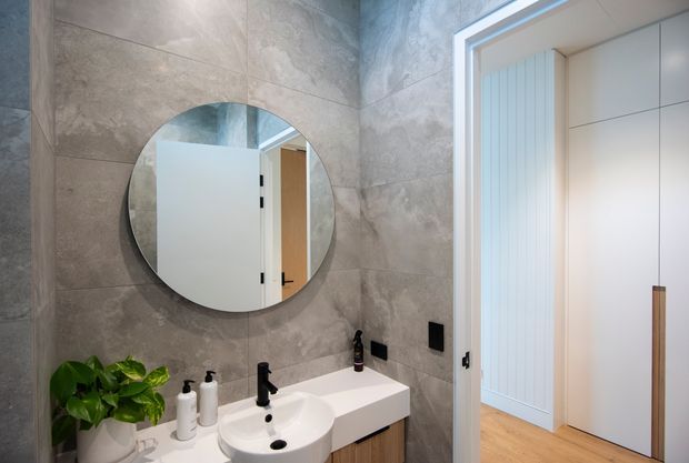 Contemporary bathroom with large grey stone-look wall tiles, a round wall-mounted mirror, a white vessel basin on a floating vanity, and black tapware and accessories. A white built-in storage unit and timber flooring are visible through the open doorway, with a potted plant adding greenery.