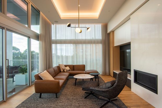 A contemporary living room with floor-to-ceiling glazing, sheer curtains, a large brown sectional sofa, lounge chairs, nesting coffee tables, timber flooring, and a dark textured rug. A built-in media wall with television and linear fireplace occupies the right side, while a recessed ceiling with concealed cove lighting and a sculptural pendant adds architectural interest. The glazed opening connects to an outdoor terrace or balcony.