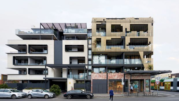 Street-level view of a contemporary multi-storey apartment and mixed-use building. The facade combines projecting balconies, extensive glazing, dark metal cladding, pale rendered surfaces, exposed concrete, and a lower-level brick commercial frontage. A tram and streetscape are visible in the foreground.