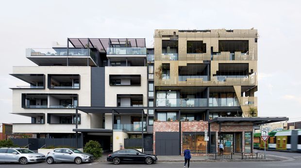 Street-facing view of a contemporary multi-storey mixed-use apartment building, with irregular projecting balconies, extensive glazing, rooftop terraces, and contrasting white rendered, dark-panelled, and weathered masonry facades. Retail or commercial tenancies occupy the ground floor, with a tram and urban streetscape visible in front.