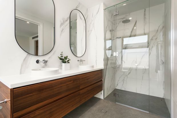 Contemporary bathroom with a long double vanity featuring dark timber drawer cabinetry and a white benchtop, two vessel basins, and rounded black-framed mirrors. The adjacent walk-in shower has clear frameless glass, chrome fixtures, large white marble-look wall tiles, and a dark grey tiled floor. The space is bright, refined, and minimally styled.