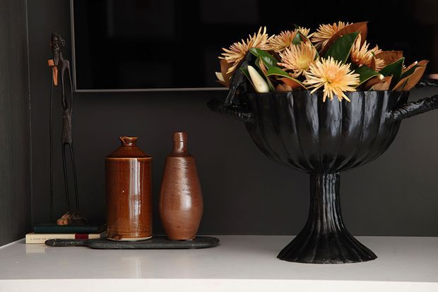 A moody styled interior vignette on a light stone or painted shelf, featuring a large black sculptural pedestal bowl filled with dried flowers and foliage, alongside two brown ceramic vessels, a small tray, books, and a slender dark figurine. A black television or artwork occupies the upper background, with dark charcoal walls creating a dramatic, refined setting.