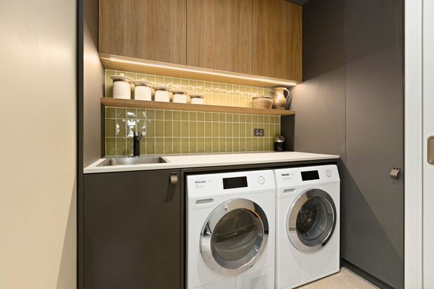 Compact, contemporary laundry room with front-loading white washer and dryer beneath a white benchtop. Dark grey cabinetry, warm timber overhead cupboards, and a glossy olive-green tiled splashback create a refined, warm-toned fit-out. Open shelving with containers and integrated under-cabinet lighting add practical storage and illumination.