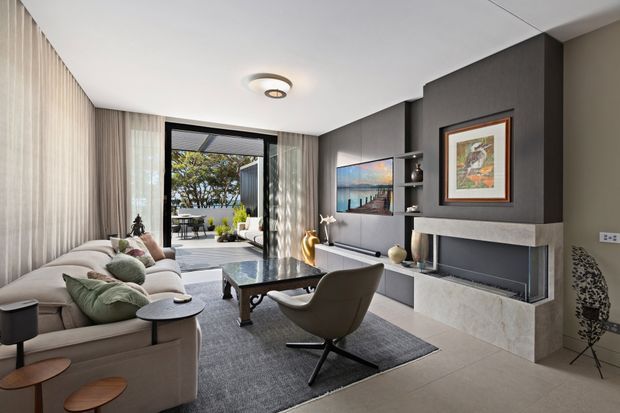 Contemporary living room with a large sectional sofa, sculptural lounge chair, dark coffee table, grey rug, and a built-in fireplace wall. The room features floor-to-ceiling sheer curtains, dark charcoal feature joinery with open shelving, pale stone fireplace cladding, tiled flooring, and a large glazed opening to an outdoor terrace. Neutral grey, cream, and black tones create a refined, comfortable interior.