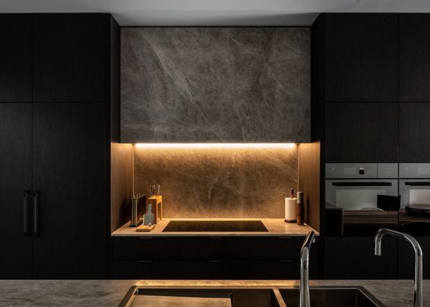 A dark, sophisticated contemporary kitchen viewed straight on. Full-height black or very dark timber cabinetry frames a central cooking zone with a large veined stone feature splashback and matching benchtop. Warm concealed LED lighting illuminates the splashback and work surface. A black induction cooktop, integrated double ovens, sink and tall chrome gooseneck tap are visible, creating a moody, luxurious atmosphere.