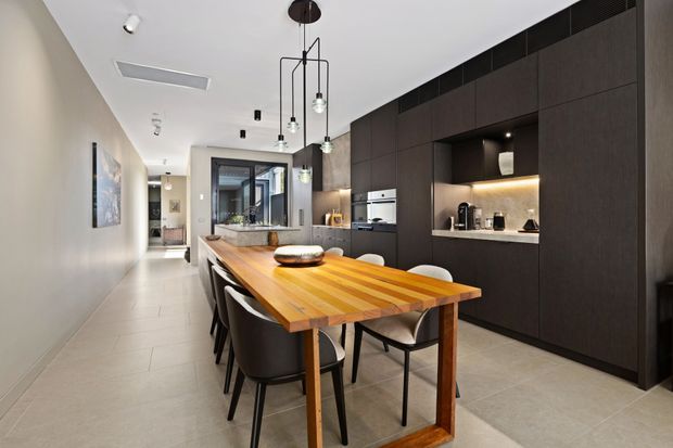 A contemporary open-plan kitchen and dining area viewed from a long circulation space. A large warm timber dining table with dark upholstered chairs sits in the foreground, while a dark full-height built-in kitchen wall includes integrated appliances, open display niches, and under-cabinet lighting. Pale large-format tiled flooring, light neutral walls, recessed and pendant lighting, and a distant glazed opening create a refined, elongated interior.
