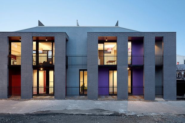 Front exterior view of a contemporary multi-storey residential building with a symmetrical facade of dark grey brick piers, recessed balconies, tall glazed openings, and black metal balustrades. Warm interior lighting and colourful red and purple walls are visible through the openings. The building has a low-pitched roof and a narrow urban frontage.