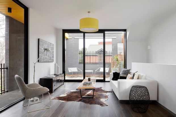 A bright contemporary living room with dark timber flooring, white walls, and a large black-framed glazed opening leading to a balcony. The room contains a white sofa, textured grey lounge chair, low timber coffee table, media console, floor lamp, patterned ottoman, and a bold yellow pendant light. A dark exposed brick wall and warm timber ceiling lining are visible at the left, while neighbouring urban buildings can be seen beyond the glazing.