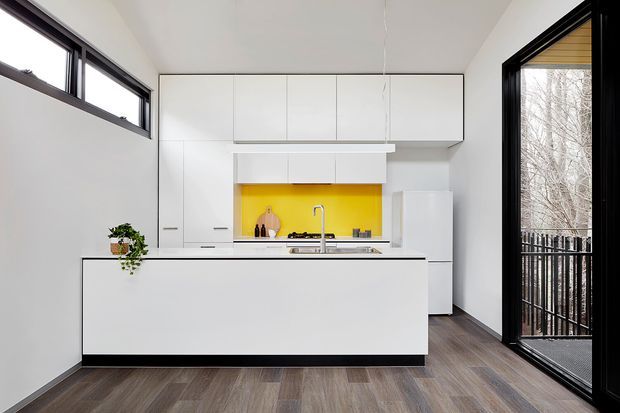 A bright, compact contemporary kitchen with extensive flat-front white cabinetry, a large white island, dark-framed windows and a full-height glazed door. A vivid yellow splashback provides a strong accent behind the stainless-steel sink and cooktop, while dark wood-look flooring adds warmth. The space opens directly to an exterior balcony or deck.