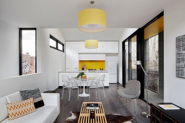 Bright, compact open-plan interior combining a living area, dining space and contemporary white kitchen. The room features dark timber flooring, white cabinetry, black-framed windows and full-height glazing, transparent dining chairs, a white sofa, a grey lounge chair, and bold yellow pendant lights with a matching yellow kitchen splashback. The space has a clean modern aesthetic with strong black-and-white contrast and warm yellow accents.