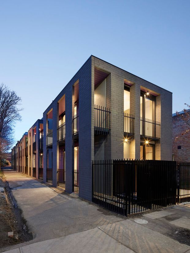 Twilight exterior view of a contemporary multi-storey residential terrace development along a narrow urban lane. The repeated townhouse facades use pale grey masonry, deep vertical window reveals, recessed balconies, dark metal balustrades and secure fencing, with warm interior and exterior lighting accentuating the elevations.