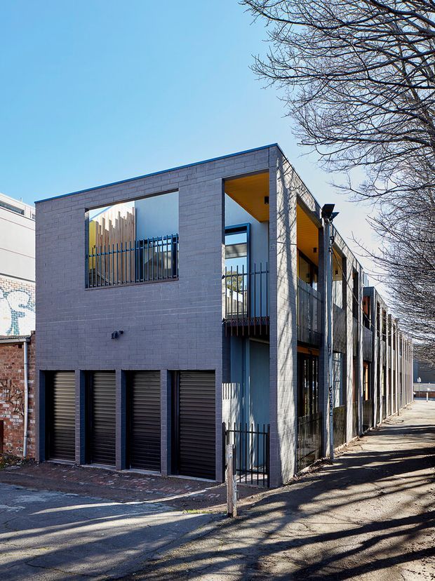 Exterior street view of a narrow, multi-storey contemporary urban building. The facade is composed of pale grey masonry walls, dark metal security grilles and roller doors, recessed balconies, and warm timber-lined soffits. A long repetitive side elevation follows the sidewalk, with mature leafless trees framing the scene.
