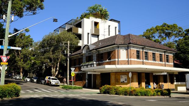 Street-level view of a corner commercial building combining a traditional brick masonry lower structure with a tiled hipped roof and a contemporary white multi-storey addition above. The building fronts a leafy urban intersection with marked pedestrian crossings, street signage, parked vehicles, and mature trees.
