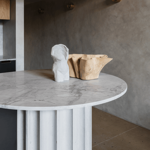 Close-up of a sculptural round stone kitchen island or counter with a fluted cylindrical base, set against a textured grey plaster wall. A white sculptural bust and a pale wood vessel sit on the benchtop, with dark bronze pendant or wall fittings visible above. The scene has a refined, moody contemporary character.