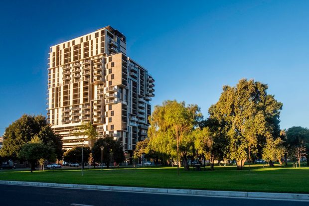 A tall contemporary multi-storey apartment building with a strongly articulated façade of balconies, vertical window bands, projecting box-like elements and light-toned cladding. The tower stands beside a broad landscaped green space with mature trees, viewed from across a paved road under clear blue sky.