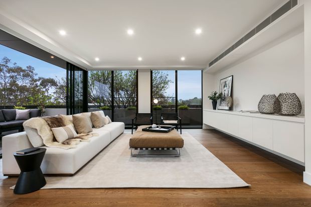 A bright, contemporary living room with a large white sectional sofa, black sculptural side table, low coffee table and upholstered ottoman arranged on a pale area rug. Floor-to-ceiling glazed sliding doors open to a leafy exterior, while a long white built-in media/storage cabinet runs along the right wall. Recessed ceiling lights, timber flooring, and a linear ceiling air-conditioning vent complete the refined minimalist interior.