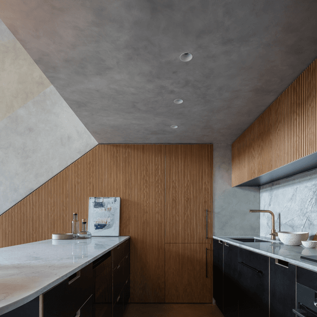 Contemporary kitchen featuring a large pale stone island, dark cabinetry, a textured grey concrete-look ceiling and walls, and warm vertical timber slat panelling across the rear wall and upper cabinetry. Integrated appliances and minimal recessed lighting contribute to a refined, material-focused interior.