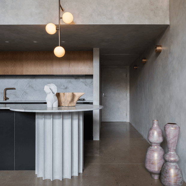 A refined contemporary kitchen and hallway interior with a dark stone island, fluted light-toned island base, black cabinetry, textured grey plaster walls, and a timber slatted feature wall. Sculptural pendant globes hang above the island, while a long corridor with subtle wall lights leads to a closed door. Metallic ceramic vessels add decorative interest in the foreground.