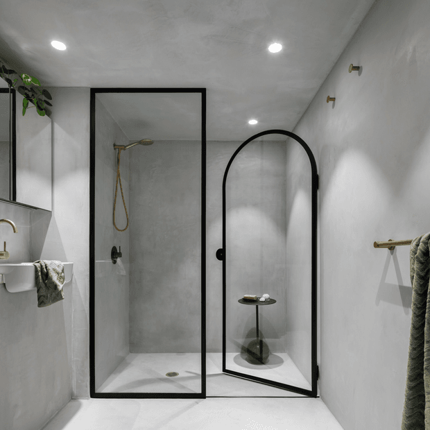 A compact contemporary bathroom with a frameless-style glass shower enclosure featuring black metal framing and a rounded glass door. The room has smooth light grey plaster or microcement-like wall and floor finishes, recessed ceiling spotlights, brass shower fittings and towel hardware, a wall-mounted basin at left, and a small black side table visible through the curved shower door. A few hanging towels and a trailing plant add texture.
