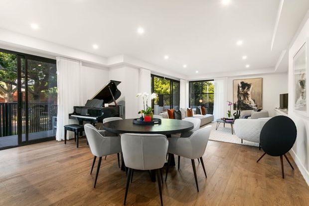 Bright, spacious open-plan living and dining room with a round dining table and upholstered chairs in the foreground, a black grand piano beside full-height glazed doors, and a contemporary lounge area beyond. The room has white walls and ceiling, recessed downlights, warm timber flooring, large black-framed windows and doors, modern artwork, and abundant natural light.