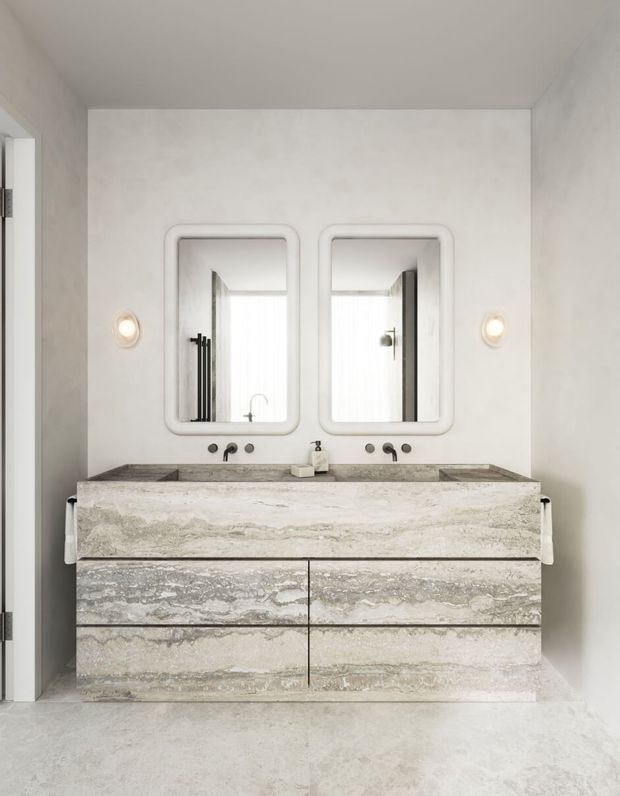 A refined minimalist bathroom featuring a floating double vanity with a thick pale natural-stone benchtop and matching stone drawer fronts. Two rounded-rectangle mirrors, wall-mounted basin taps, sculptural wall lights, and pale grey stone or plaster surfaces create a calm, monochromatic interior. Reflections reveal a shower area behind the camera-facing wall.