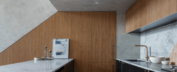 Contemporary kitchen interior featuring dark lower cabinetry, a pale stone island benchtop, a marble-look splashback, warm vertical timber wall panelling, and matching timber slatted overhead cabinetry. Minimal decor and integrated appliances create a refined, modern fit-out.
