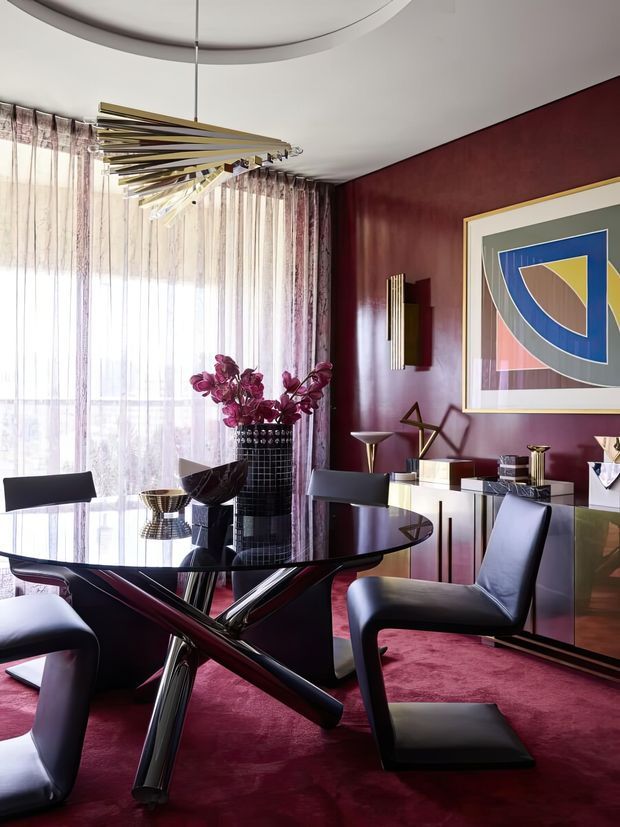 A dramatic, richly styled dining room with a glossy black dining table, sculptural chrome and dark legs, and black leather dining chairs. Full-height sheer curtains filter strong daylight along one wall, while deep burgundy painted walls and carpet create a luxurious backdrop. A long sideboard, abstract artwork, decorative objects, and a sculptural linear pendant light complete the sophisticated interior.