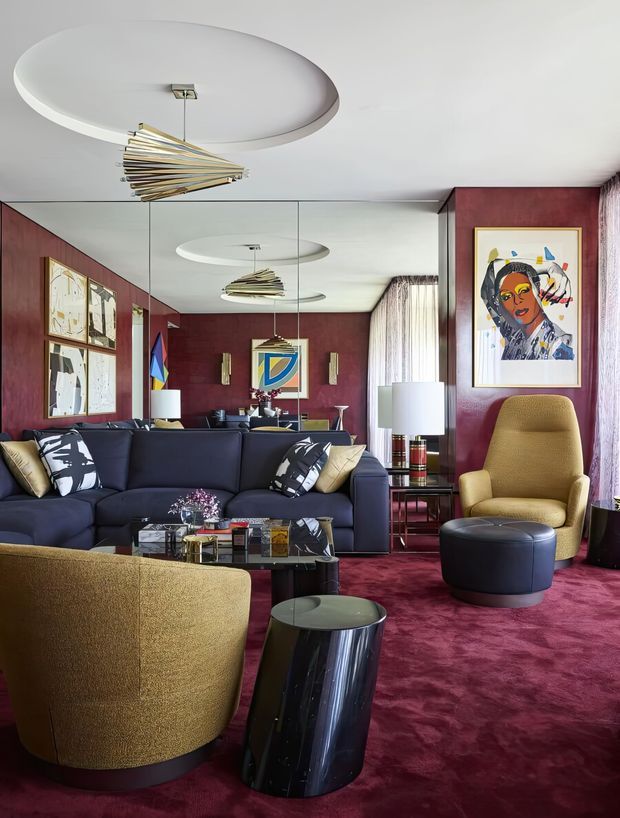 A richly layered living room with deep burgundy walls and carpet, a large mirrored feature wall, navy sectional sofa, mustard boucle lounge chairs, black glass coffee tables, framed artwork, and sculptural brass-and-white ceiling lights. The space has a dramatic, colourful, retro-luxe character with strong geometric and mid-century-inspired furnishings.