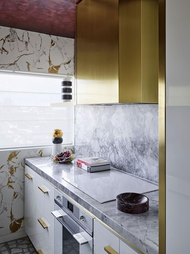 A compact, highly detailed contemporary kitchen featuring white and grey veined marble-look surfaces across the benchtop and splashback, white cabinetry with slim brass-gold handles, tall olive-bronze timber-toned cabinetry, integrated stainless-steel appliances, and a dark reddish-brown textured ceiling. A large window provides bright natural light.