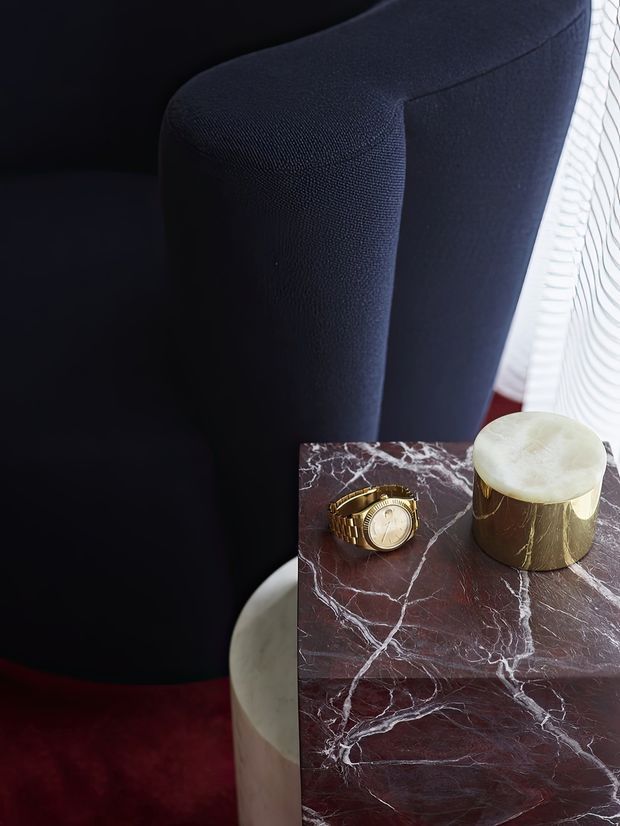 Close-up interior detail featuring a dark navy upholstered lounge chair beside a richly veined burgundy marble side table. The table holds a gold wristwatch and a cylindrical metallic vessel with a pale stone-like top. A deep red rug and a softly patterned sheer curtain add layered colour and texture. The composition is moody, luxurious, and editorial, with no broader room context visible.