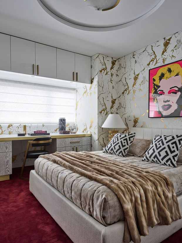 A glamorous bedroom with a large upholstered bed dressed in taupe and brown textured bedding, bold black-and-white geometric cushions, and a vivid pop-art portrait. The room features white cabinetry with brass-toned handles, a built-in desk and vanity area, a dramatic white marble-look wall finish with gold veining, deep red carpet, and a sculptural circular ceiling light.