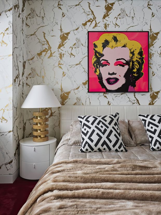 A glamorous bedroom with a padded upholstered bed, layered beige bedding, bold black-and-white geometric cushions, and a round white bedside table. The walls are clad in dramatic white marble-look material with gold veining, complemented by a vivid pop-art portrait above the bed. A sculptural gold table lamp and deep burgundy carpet add luxurious contrast.