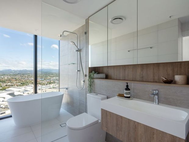 Bright contemporary bathroom with a freestanding white bathtub beside floor-to-ceiling glazing, a frameless glass shower, wall-hung toilet, and floating timber-look vanity with an integrated white basin. Mirrored wall cabinets, pale tiled surfaces, and a city-and-hills outlook create an airy high-rise setting.