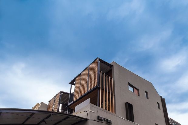 Low-angle exterior view of a contemporary multi-storey residential building. The facade combines smooth dark grey rendered walls with prominent warm timber-look horizontal screening and vertical timber fins, recessed balconies or openings, and a projecting metal-framed carport roof. The building is set against a blue sky.